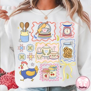 May include: White sweatshirt with a colourful illustration of baking items. The design includes a rolling pin, tea set, scales, jars of cookies and jam, wooden spoons, and a chicken. The design is framed with decorative borders.