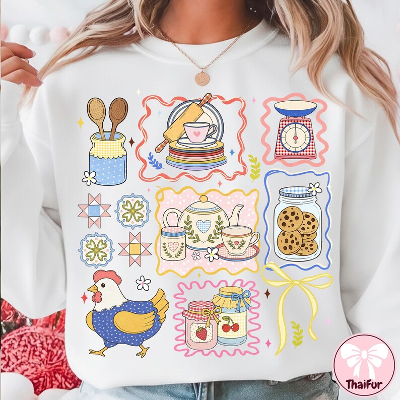 May include: White sweatshirt with a colourful illustration of baking items. The design includes a rolling pin, tea set, scales, jars of cookies and jam, wooden spoons, and a chicken. The design is framed with decorative borders.