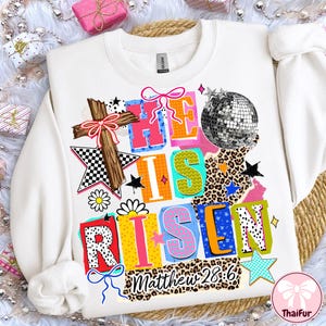 Pode incluir: Camisola branca com um gráfico colorido que diz "HE IS RISEN" com uma cruz, uma bola de discoteca e estrelas. O texto "Matthew 28:6" também está incluído. A camisola tem gola redonda.