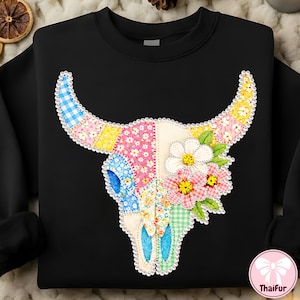 Puede incluir: Sudadera negra con un diseño de calavera de toro en patchwork. La calavera presenta patrones florales y de cuadros en tonos rosa, azul, amarillo y verde. El diseño incluye flores blancas en forma de margarita.