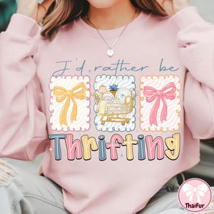 May include: A light pink sweatshirt with the text "I'd rather be thrifting." The graphic includes a yellow bow, a shopping cart with dishes, and a pink bow. The word "Thrifting" is in a colorful, bubble font.