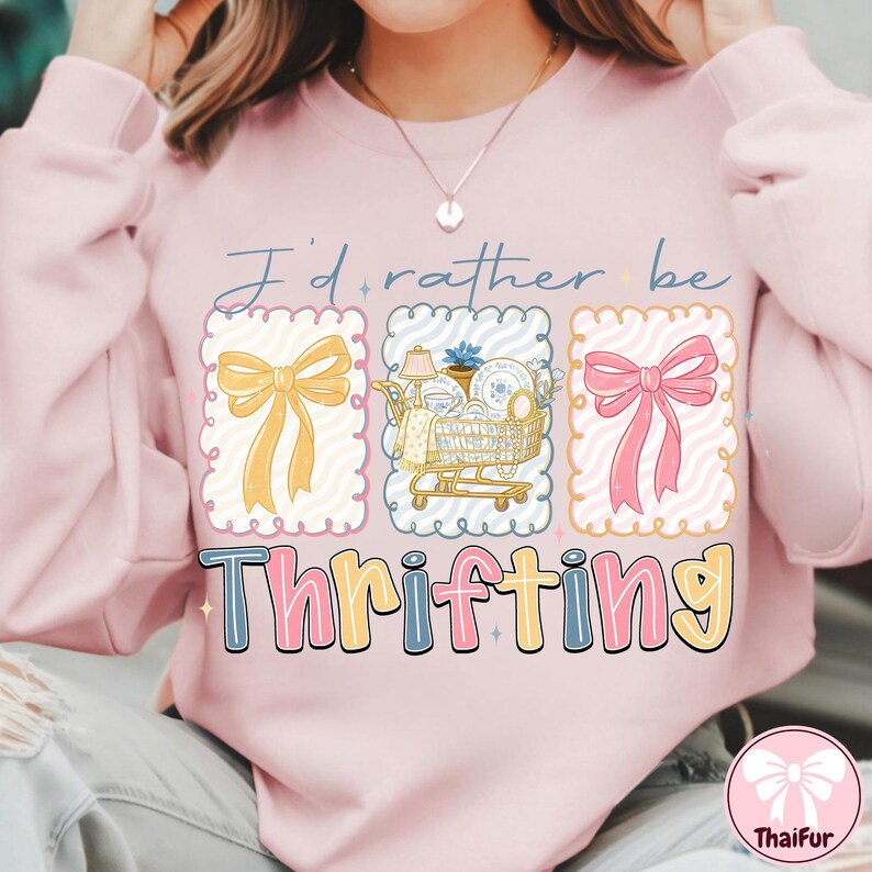 May include: A light pink sweatshirt with the text "I'd rather be thrifting." The graphic includes a yellow bow, a shopping cart with dishes, and a pink bow. The word "Thrifting" is in a colorful, bubble font.