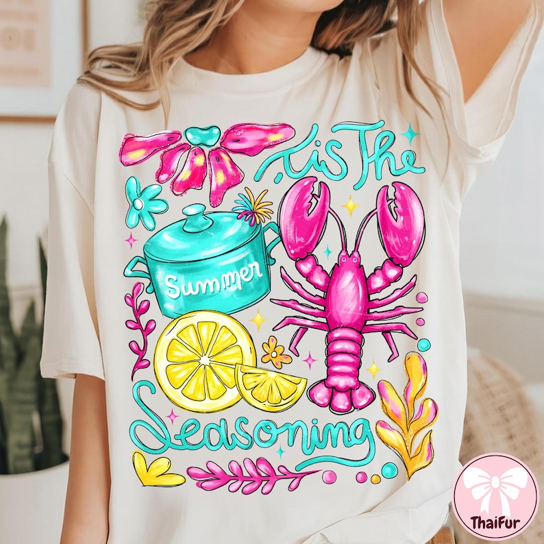 May include: Cream-colored t-shirt with a colorful graphic. The design features a pink lobster, a blue pot with the word "Summer", and a lemon slice. The text "'Tis The Seasoning" is written in turquoise.