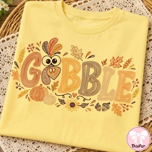 May include: A light yellow t-shirt featuring a graphic print with the text GOBBLE in large, multi-colored, line-art style letters. The design includes a turkey face, pumpkins, autumn leaves, sunflowers, and acorns in shades of orange, brown, and gold.