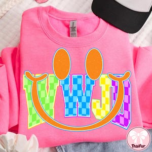 May include: A pink sweatshirt with a smiley face design. The design includes orange oval eyes, a colorful smile, and the letters "WWJ" in a checkered pattern. The sweatshirt is made of a soft material and is suitable for casual wear.