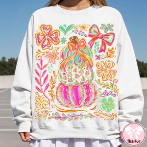 May include: White sweatshirt featuring a colorful stacked pumpkin design with bows and floral accents. The pumpkins are pink, green, and orange, with a brown stem and a bow on top. Surrounding the pumpkins are flowers and leaves in shades of pink, orange, and green.
