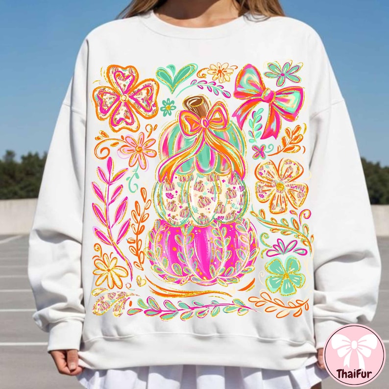 May include: White sweatshirt featuring a colorful stacked pumpkin design with bows and floral accents. The pumpkins are pink, green, and orange, with a brown stem and a bow on top. Surrounding the pumpkins are flowers and leaves in shades of pink, orange, and green.