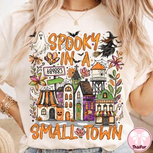 May include: A cream-colored t-shirt featuring a colorful Halloween-themed graphic with the text "SPOOKY IN A SMALL TOWN". The design includes a witch on a broomstick, ghosts, a water tower, various buildings, spiderwebs, flowers, and a sign reading 689883.