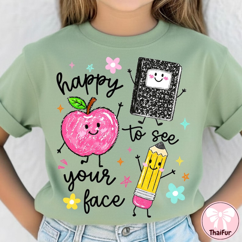 May include: A sage green t-shirt featuring a graphic print of a pink apple, a black and white composition notebook, and a yellow pencil, all with smiling faces. The text reads "happy to see your face" in black script, surrounded by small colorful stars and flowers.