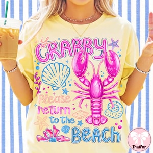 Puede incluir: Camiseta amarilla con un diseño gráfico rosa y azul. El diseño incluye una langosta rosa, conchas marinas y el texto "If Crabby Please Return to the Beach". La camiseta tiene mangas cortas y un corte holgado.