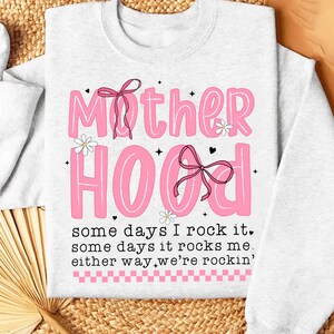 May include: A white sweatshirt with a pink graphic that says "Mother Hood" with pink bows and flowers. The text below says "some days I rock it, some days it rocks me, either way we're rockin'."