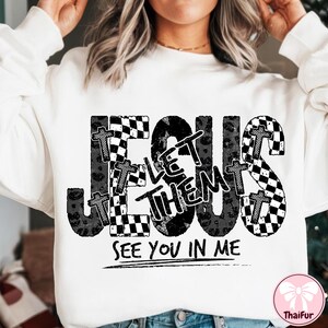 May include: White sweatshirt with a graphic design featuring the word "JESUS" in large, distressed black letters. The design includes the words "LET THEM" and "SEE YOU IN ME". Crosses and checkered patterns are incorporated.