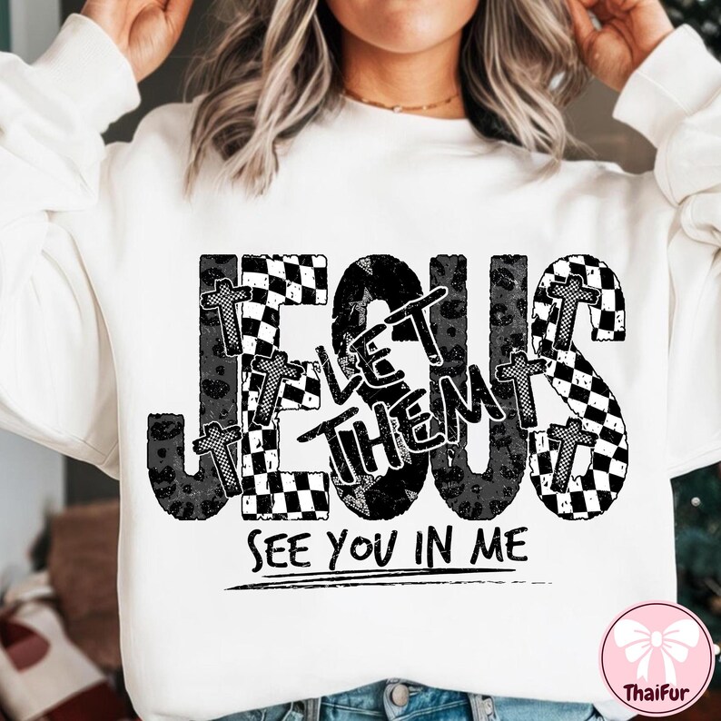 May include: White sweatshirt with a graphic design featuring the word "JESUS" in large, distressed black letters. The design includes the words "LET THEM" and "SEE YOU IN ME". Crosses and checkered patterns are incorporated.