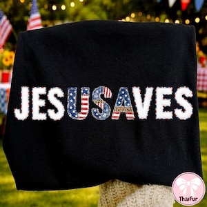 May include: Black sweatshirt with the text "JESUS SAVES" in white, with the letters "USA" in a patchwork design featuring the American flag and leopard print. The background includes American flags and festive decorations.