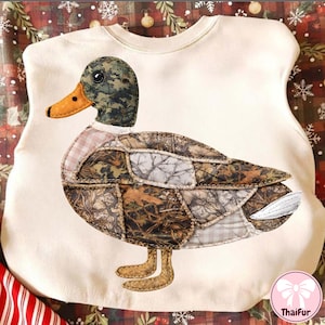 May include: Cream-colored sweatshirt featuring a large, appliqued duck. The duck has a camouflage patterned body, a green and brown head, and an orange beak. The duck's feet are tan, and the sweatshirt has a round neckline.