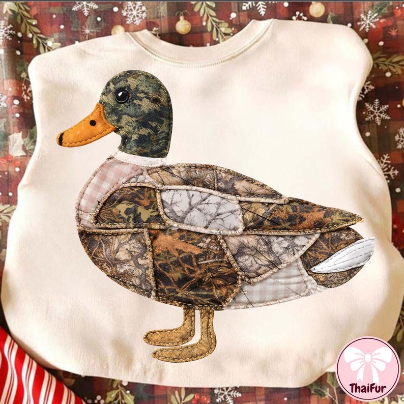 May include: Cream-colored sweatshirt featuring a large, appliqued duck. The duck has a camouflage patterned body, a green and brown head, and an orange beak. The duck's feet are tan, and the sweatshirt has a round neckline.