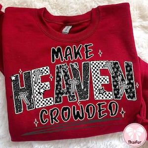 May include: A red sweatshirt with the phrase "MAKE HEAVEN CROWDED" in a black and white design. The word "HEAVEN" features stars, crosses, and a checkered pattern. The sweatshirt has a crew neck and long sleeves.
