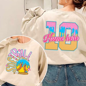 May include: A beige sweatshirt with a pink and blue ice cream cone design on the back. The design includes the number 10 and the text "Name here". The front of the sweatshirt has a colorful graphic of a softball with the word "Softball" written around it.