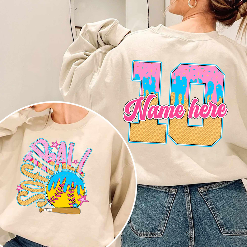 May include: A beige sweatshirt with a pink and blue ice cream cone design on the back. The design includes the number 10 and the text "Name here". The front of the sweatshirt has a colorful graphic of a softball with the word "Softball" written around it.