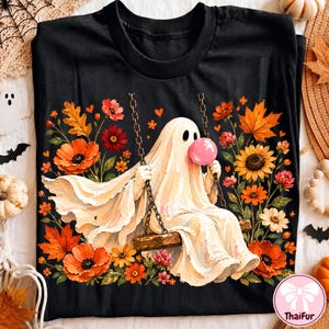 May include: A black t-shirt featuring a graphic of a white ghost sitting on a wooden swing, blowing a pink bubble. The ghost is surrounded by colorful autumn flowers and leaves, including orange poppies, sunflowers, and red maple leaves.