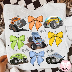 K&ouml;nnte beinhalten: Ein hellgraues Sweatshirt mit bunten Cartoon-Monstertrucks und Schleifen. Die Monstertrucks sind in Orange, Gelb, Gr&uuml;n, Blau und Grau gehalten und haben gro&szlig;e Reifen und aggressive Gesichtsausdr&uuml;cke. Die Schleifen sind orange, gr&uuml;n, gelb und blau.