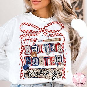 Può includere: Felpa bianca con grafica a tema baseball. Il design include le parole "Hey Batter Batter Swing" in rosso, bianco e blu, insieme a illustrazioni di attrezzature da baseball. Un nastro a scacchi rosso e bianco incornicia il testo.