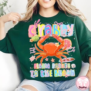 May include: Dark green sweatshirt featuring a crab graphic and the text "If Crabby, Please Return To The Beach". The crab is orange with yellow details, and the text is in bright colours. The sweatshirt has a crew neck.