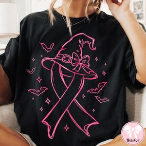 Könnte beinhalten: Ein schwarzes T-Shirt mit einem leuchtend pinkfarbenen Grafikdruck, der eine Schleife zur Sensibilisierung zeigt, die von einem Hexenhut mit Schleife gekrönt wird. Das Motiv ist von kleinen pinken Fledermäusen und vierzackigen Sternen umgeben.