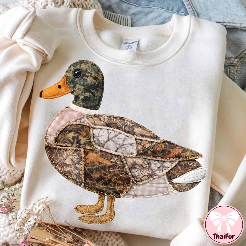 May include: Cream-colored sweatshirt featuring a large, appliqued duck design. The duck has a camouflage patterned head and body, with a yellow-orange beak and brown feet. The design is made of various fabric patches.