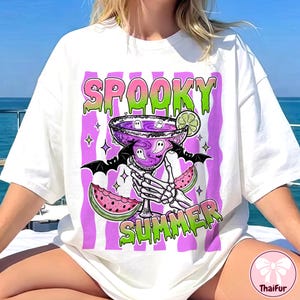 Puede incluir: Camiseta blanca con un gráfico colorido que dice "Spooky Summer". El diseño presenta un cóctel morado con detalles de fantasmas, alas de murciélago, rodajas de sandía y una mano de esqueleto. El texto es en verde y rosa.