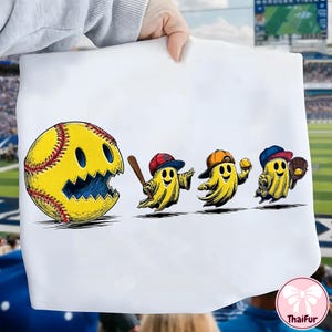 Puede incluir: Tela blanca con un diseño de dibujos animados. Una pelota de softbol amarilla con una cara sonriente está junto a tres figuras fantasmales jugando béisbol. Las figuras llevan gorras de béisbol y sostienen un bate, una pelota y un guante.