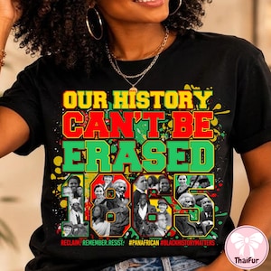 Op de afbeelding: Een zwart t-shirt met de tekst "OUR HISTORY CAN'T BE ERASED" in gele, rode en groene blokletters. Het ontwerp bevat een collage van historische figuren en verfspatten in de kleuren geel, rood en groen.