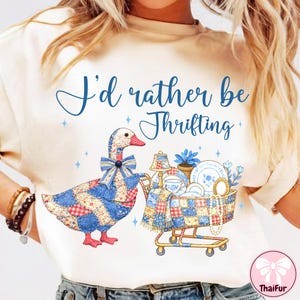 以下が含まれることがあります： 「I'd rather be Thrifting」の青い文字がプリントされたクリーム色のTシャツ。パッチワークのガチョウと、お皿やランプなどのヴィンテージ品でいっぱいのショッピングカートが描かれています。