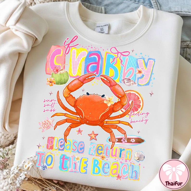 May include: Cream-coloured sweatshirt with a vibrant graphic. It features a crab, the text "If Crabby Please Return To The Beach", and beach-themed illustrations. The design includes a coconut, a slice of grapefruit, and flowers.