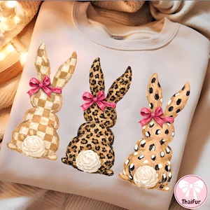 May include: Cream-colored sweatshirt featuring three decorative bunny figures. The bunnies have unique patterns: checkered, leopard print, and spotted. Each bunny has a pink bow. The ThaiFur logo is in the bottom right corner.