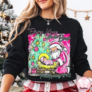 May include: Black sweatshirt with a pink and green graphic featuring a Christmas tree, Santa Claus, and baby Jesus. The text "Every Knee Shall Bow" and "Philippians 2:10" are also displayed on the sweatshirt.