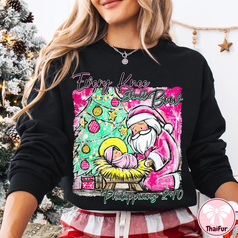 May include: Black sweatshirt with a pink and green graphic featuring a Christmas tree, Santa Claus, and baby Jesus. The text "Every Knee Shall Bow" and "Philippians 2:10" are also displayed on the sweatshirt.