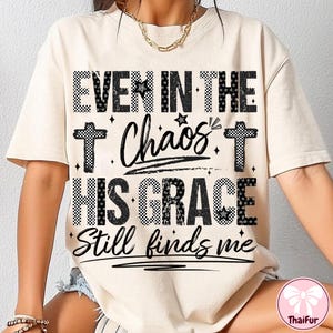 May include: Cream-colored t-shirt with the text "EVEN IN THE Chaos HIS GRACE Still finds me" in black. The text includes crosses and stars. The shirt is a casual style.