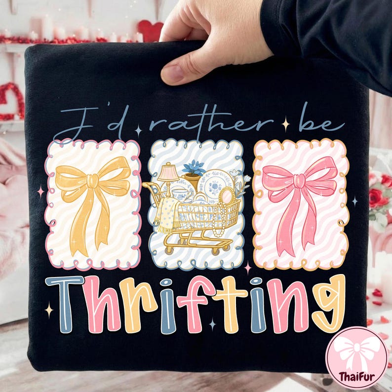 May include: Black t-shirt with the text "I'd rather be thrifting." The design includes three decorative frames with a yellow bow, a shopping cart filled with vintage items, and a pink bow. The word "Thrifting" is in colorful letters.
