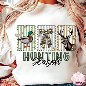 Puede incluir: Sudadera blanca con un dise&ntilde;o de rayas verdes y blancas con un pato, un lazo de camuflaje y un ciervo. Las palabras "HUNTING Season" est&aacute;n impresas en verde y negro.