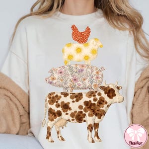 May include: Cream-colored t-shirt featuring a stacked farm animal design. A cow with brown spots forms the base, topped by a pig with floral patterns, a sheep with sun designs, and a chicken. The ThaiFur logo is visible.