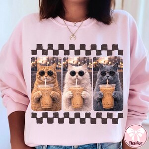 May include: Pink sweatshirt featuring three cats wearing sunglasses and drinking iced coffee. The cats are in individual frames with a brown and white checkered border. The sweatshirt has a crew neck and long sleeves.