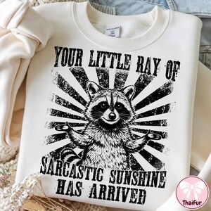 May include: Cream-colored sweatshirt with a black and white graphic of a raccoon with outstretched arms. The text reads "YOUR LITTLE RAY OF SARCASTIC SUNSHINE HAS ARRIVED" in a distressed font. The design features a sunburst pattern.