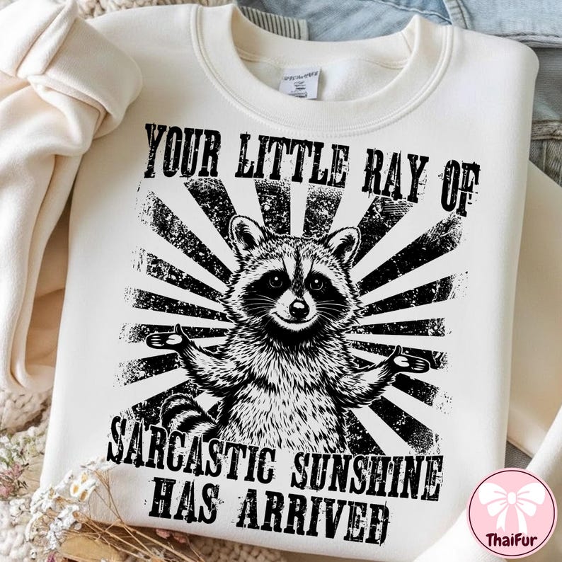 May include: Cream-colored sweatshirt with a black and white graphic of a raccoon with outstretched arms. The text reads "YOUR LITTLE RAY OF SARCASTIC SUNSHINE HAS ARRIVED" in a distressed font. The design features a sunburst pattern.