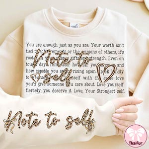 May include: Cream-colored sweatshirt with the text "Note to Self" in a leopard print design. The sweatshirt has a crew neck and long sleeves. The text "You are enough just as you are" is printed on the front.