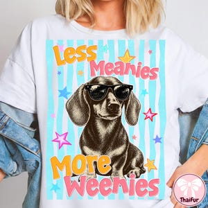May include: White t-shirt featuring a dachshund wearing sunglasses, set against a light blue striped background with colorful stars. The text reads "Less Meanies More Weenies" in orange and pink lettering. A fun, playful design.