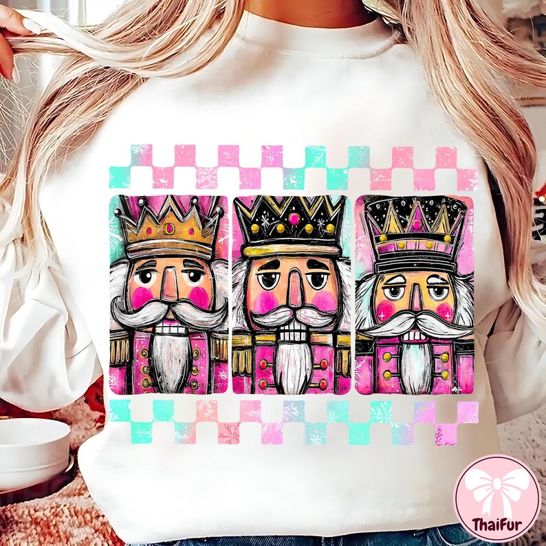 K&ouml;nnte beinhalten: Wei&szlig;es Sweatshirt mit drei farbenfrohen Nussknacker-Illustrationen. Jeder Nussknacker hat eine Krone, rosa Wangen und einen langen wei&szlig;en Schnurrbart. Das Design beinhaltet ein rosa und t&uuml;rkisfarbenes Schachbrettmuster.