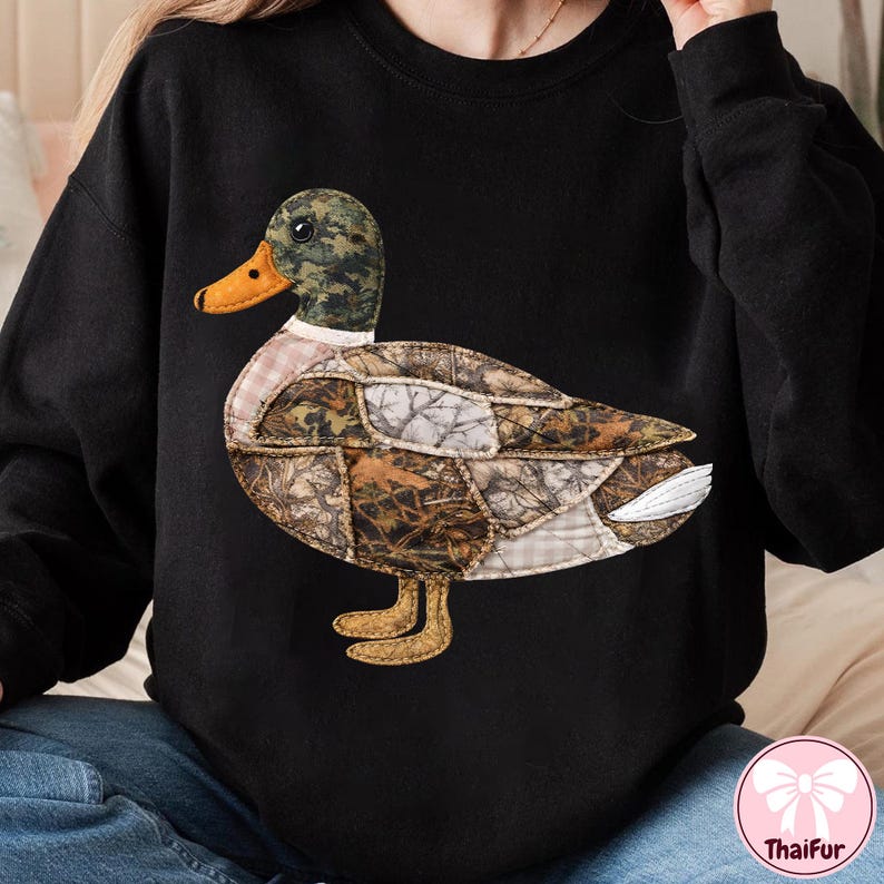 May include: Black sweatshirt featuring a large, appliqued duck design. The duck has a camouflage patterned head, a yellow beak, and a body composed of various fabric patches in shades of brown, green, and white. The duck's feet are tan.