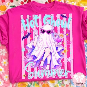 Puede incluir: Una camiseta rosa intenso con un diseño de un fantasma con gafas de sol y un cóctel. El texto "Hot Ghoul Summer" es azul claro. El fantasma hace el signo de la paz. También aparecen estrellas y murciélagos.