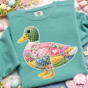 May include: Teal sweatshirt with a patchwork duck design. The duck has a green, checkered head, a yellow beak, and a body made of floral and patterned fabrics. The sweatshirt has a crew neck and long sleeves, with the brand name "Comfort Crumbs" on the tag.